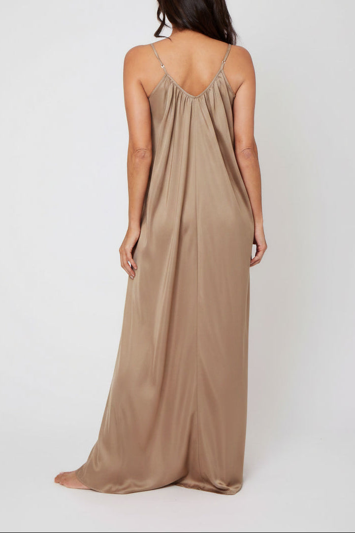 A woman viewed from the back wearing a floor-length silk dress in a warm taupe-beige color, standing against a plain light gray studio background.