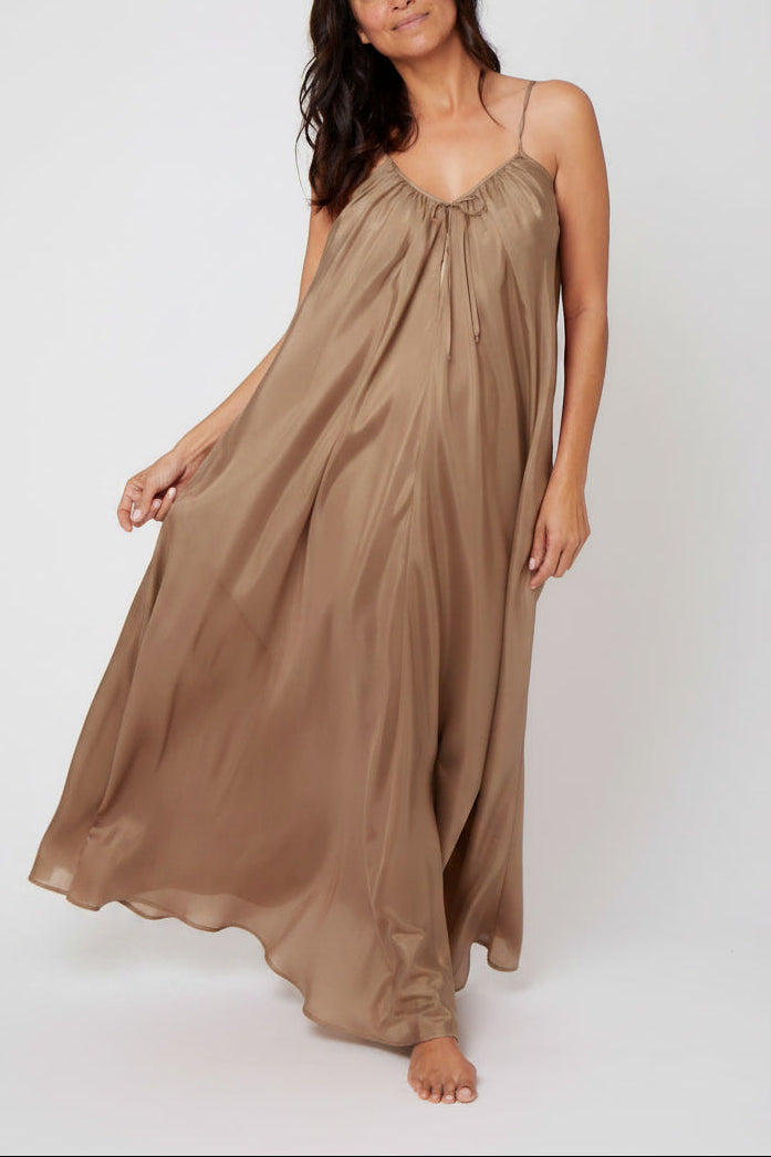 A woman modeling a flowing, sleeveless maxi silk dress in a warm taupe-brown color, standing against a plain light gray studio background.