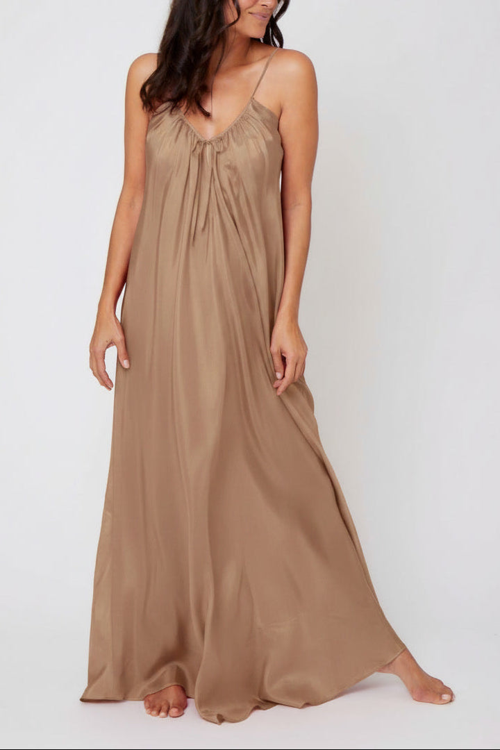 A woman modeling a floor-length silk dress in a warm taupe-brown color, standing against a plain light gray studio background.