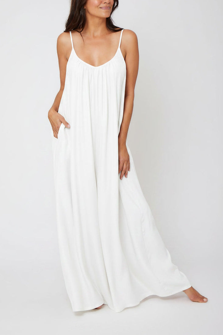 A woman modeling a white silk jumpsuit dress with thin spaghetti straps and side pockets, standing against a seamless light gray studio background.
