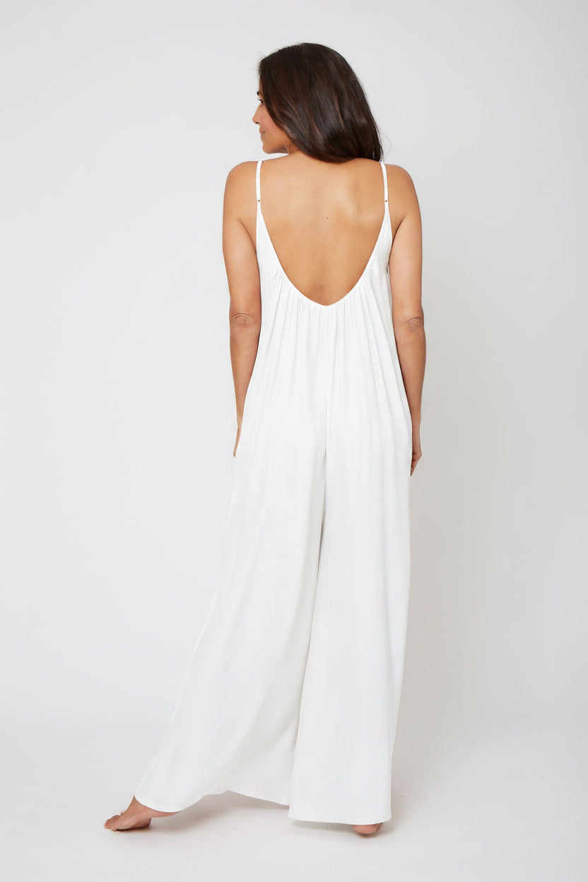 A woman viewed from behind wearing a white sleeveless jumpsuit with thin straps, a deep open back, and wide legs, standing against a plain light gray studio background.